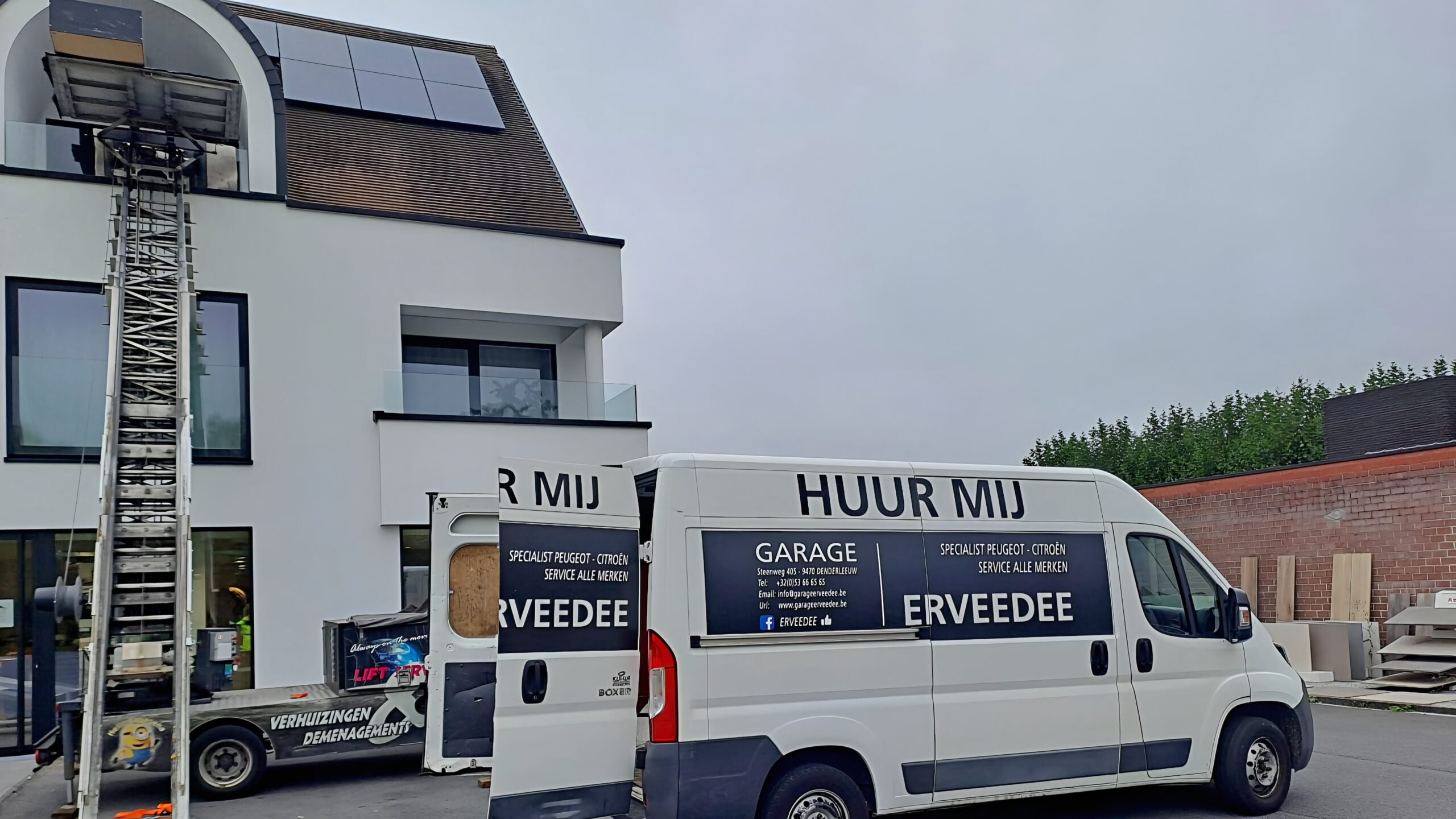 Lift de déménagement installé devant une maison moderne blanche, camionnette de déménagement garée à côté