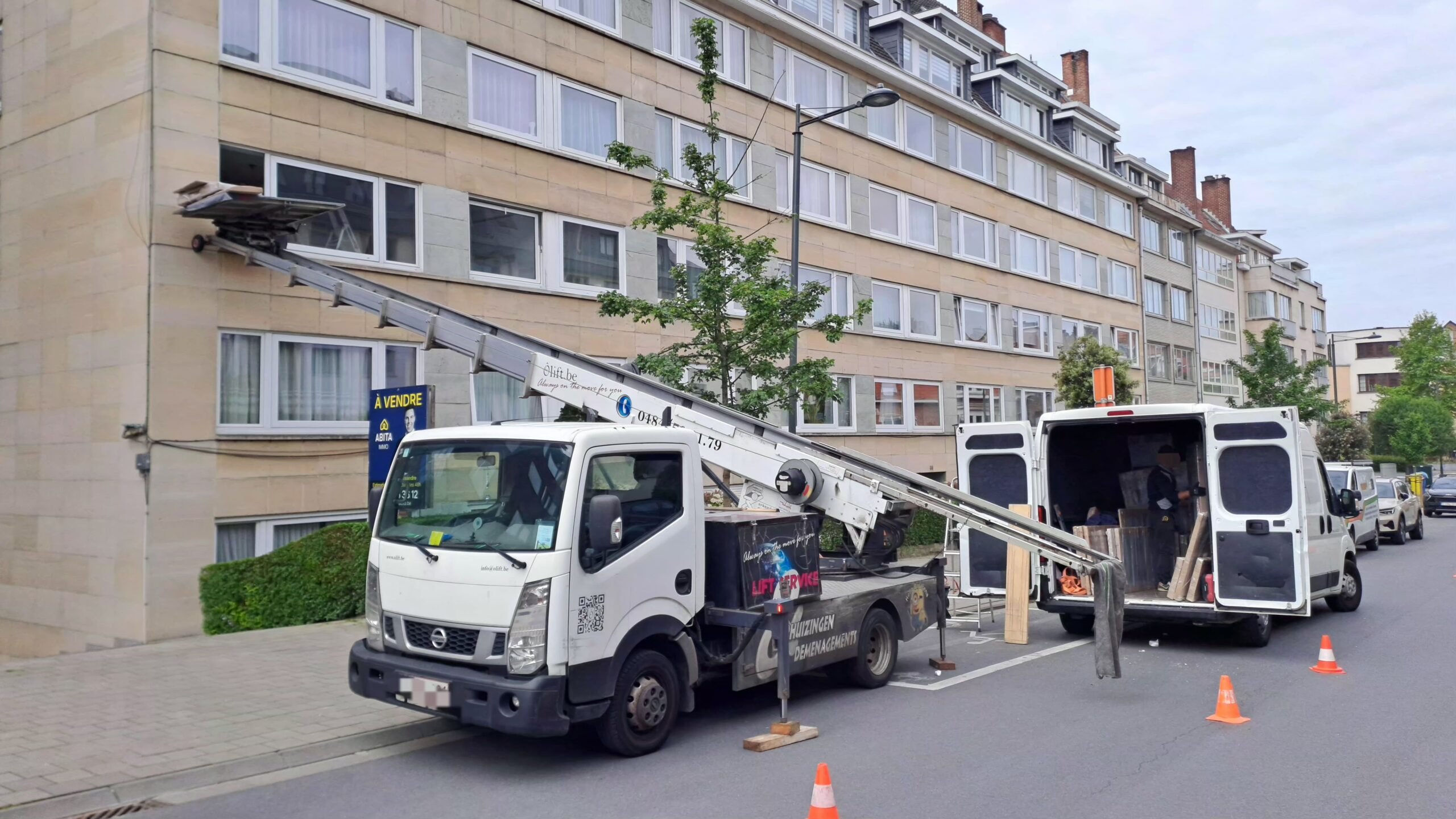 Lift de déménagement déployé vers un immeuble de logements, camionnette et cônes dans une rue résidentielle
